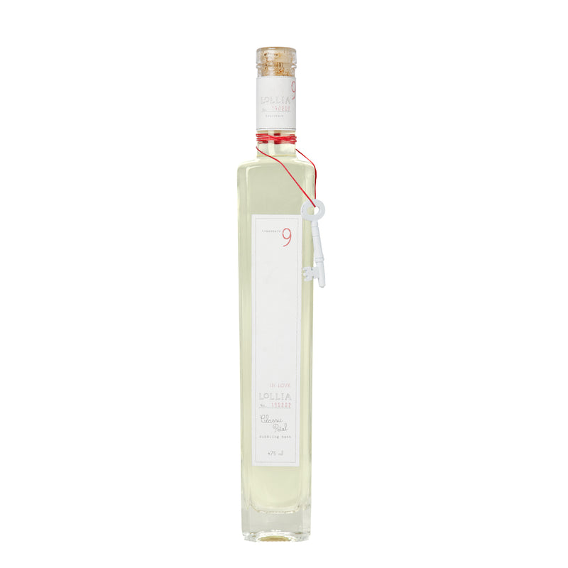 A tall, narrow bottle of Margot Elena Lollia In Love Classic Petal Bubble Bath with a clear, light yellow liquid inside. The bottle features a white label and a red seal at the neck with a string attached.
