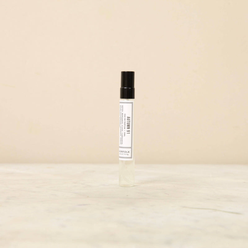 A small glass spray bottle with a black cap stands in the center on a light beige background. The label is white with black text and indicates that it contains Norfolk Natural Living Parfum - Days of Autumn 91 10ml.