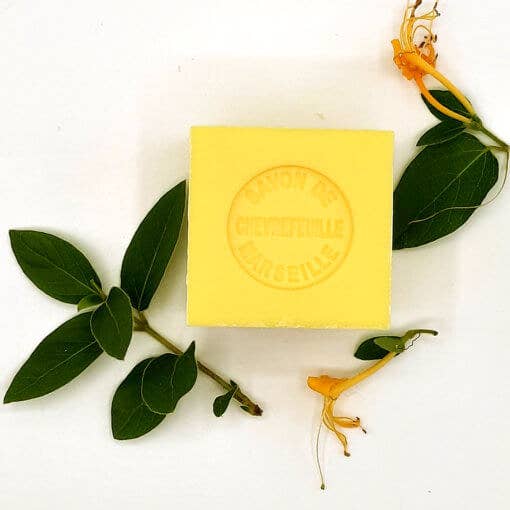 A yellow Senteurs De France Marseille Honeysuckle Cube soap bar with embossed text, surrounded by green leaves and small yellow flowers on a white background.