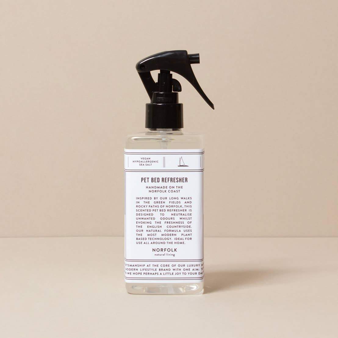 A spray bottle of Norfolk Natural Living Coastal Pet Bed Refresher, with natural ingredients, featuring a white label and black text with a trigger spray top, displayed against a light beige background.