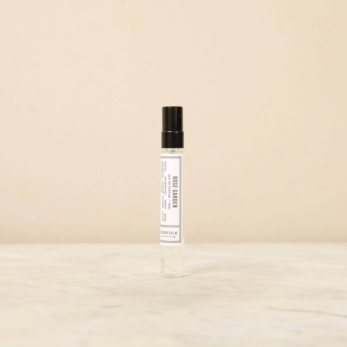 A small, cylindrical glass spray bottle with a black cap sits on a light-colored marble surface against a beige background. The bottle has a white label with black text that reads "Norfolk Natural Living Parfum - Rose Garden 10ml" and "Norfolk Natural Living," promising a long-lasting fragrance.