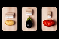 Three cutting boards from the Andrée Jardin "Canot" series, each with an Andrée Jardin "Canot" vegetable brush and a different vegetable: a potato, an eggplant, and a tomato, arranged against a black background.