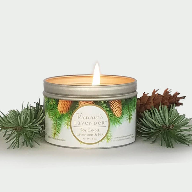 A clean-burning soy candle labeled "Victoria's Lavender - Lavender & Fir Soy Candle" in a glass jar. Surrounding the candle are pine branches and cones on a white background.