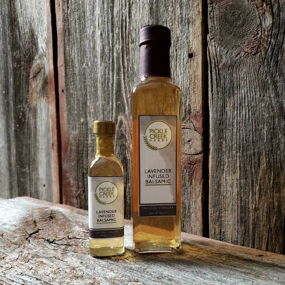 Two bottles of Pickle Creek Herbs Lavender Infused Balsamic Vinegar, one small and one tall, labeled "lavender infused," standing on a wooden surface against a rustic wooden backdrop.
