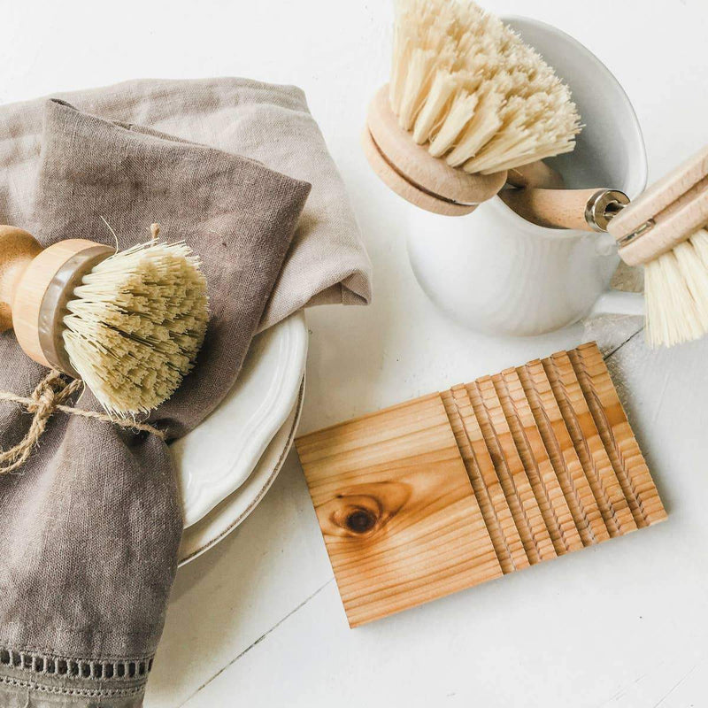A neatly arranged set of eco-friendly kitchen cleaning tools, including wooden brushes and a gray cloth on a Z&Co. Cedar Wood Block Soap Holder, on a white surface with a natural wood accent.