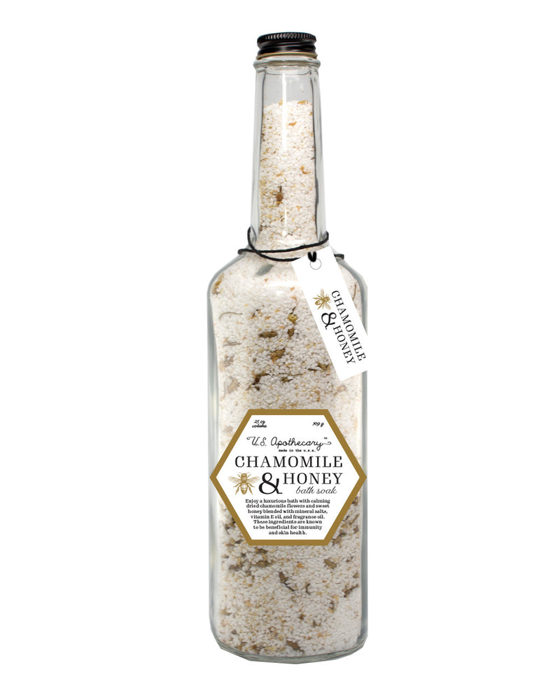 A clear glass bottle filled with U.S. Apothecary Chamomile & Honey Bath Soak Salt, labeled "all natural chamomile & honey bath salts" and sealed with a cork, featuring hanging tags detailing the product as a luxurious