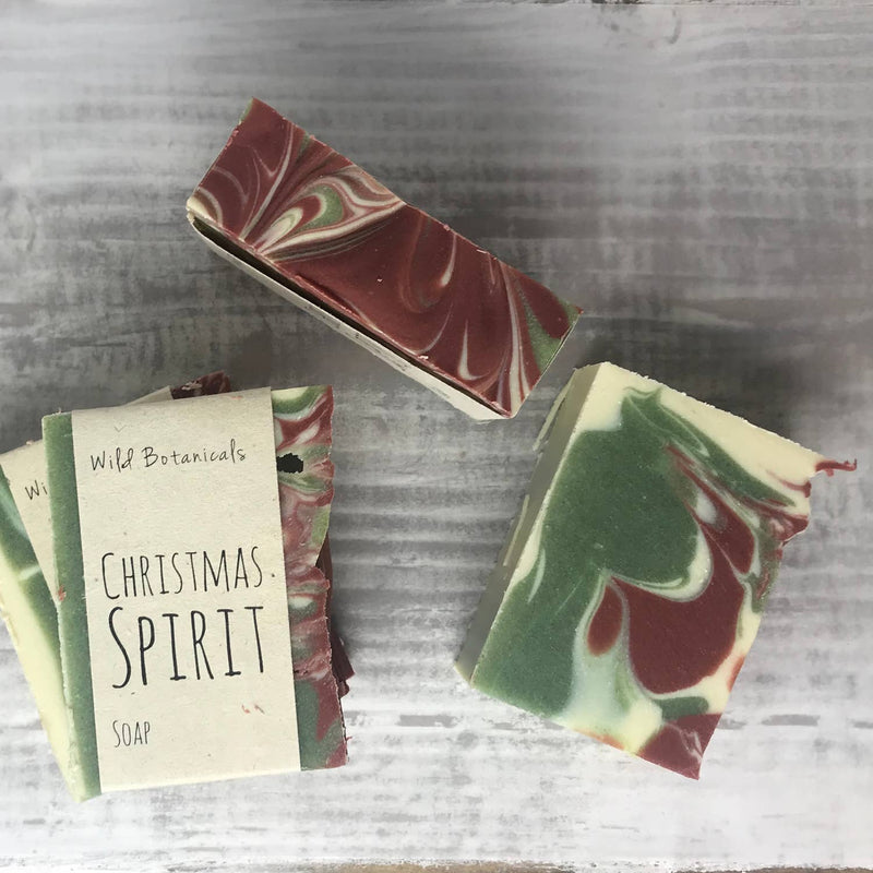 Handmade scented soap bars labeled "Wild Botanicals Christmas Spirit Soap," featuring red, green, and white swirl patterns, displayed on a wooden background.