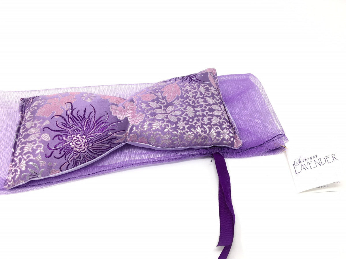 A Sonoma Lavender eye pillow, designed to soothe tired eyes, featuring floral patterns in purple with a silver border, tied with a ribbon, positioned next to a "genuine lavender" label on a.