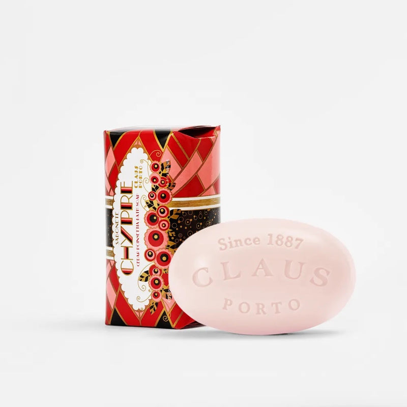 A Claus Porto 1887 soap bar infused with shea oil next to its ornate red and black packaging, featuring elaborate designs and the text "since 1887" on the soap. The background is plain.