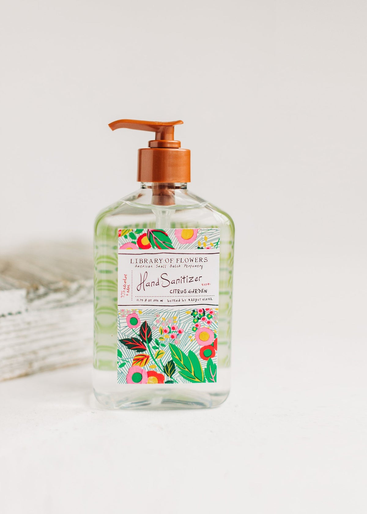 A clear bottle of Margot Elena's Library of Flowers Citrus Garden Hand Sanitizer Gel with a floral and fruit design, labeled "citrus garden," placed on a white surface against a white background.