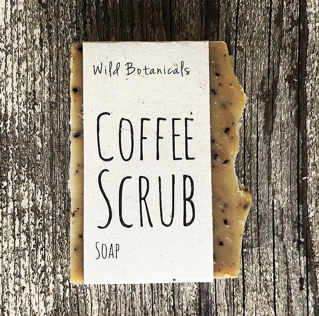 A bar of Wild Botanicals Coffee Scrub Soap, with visible organic coffee grounds, rests on a rustic wooden surface. The label is simple and features clear black text.