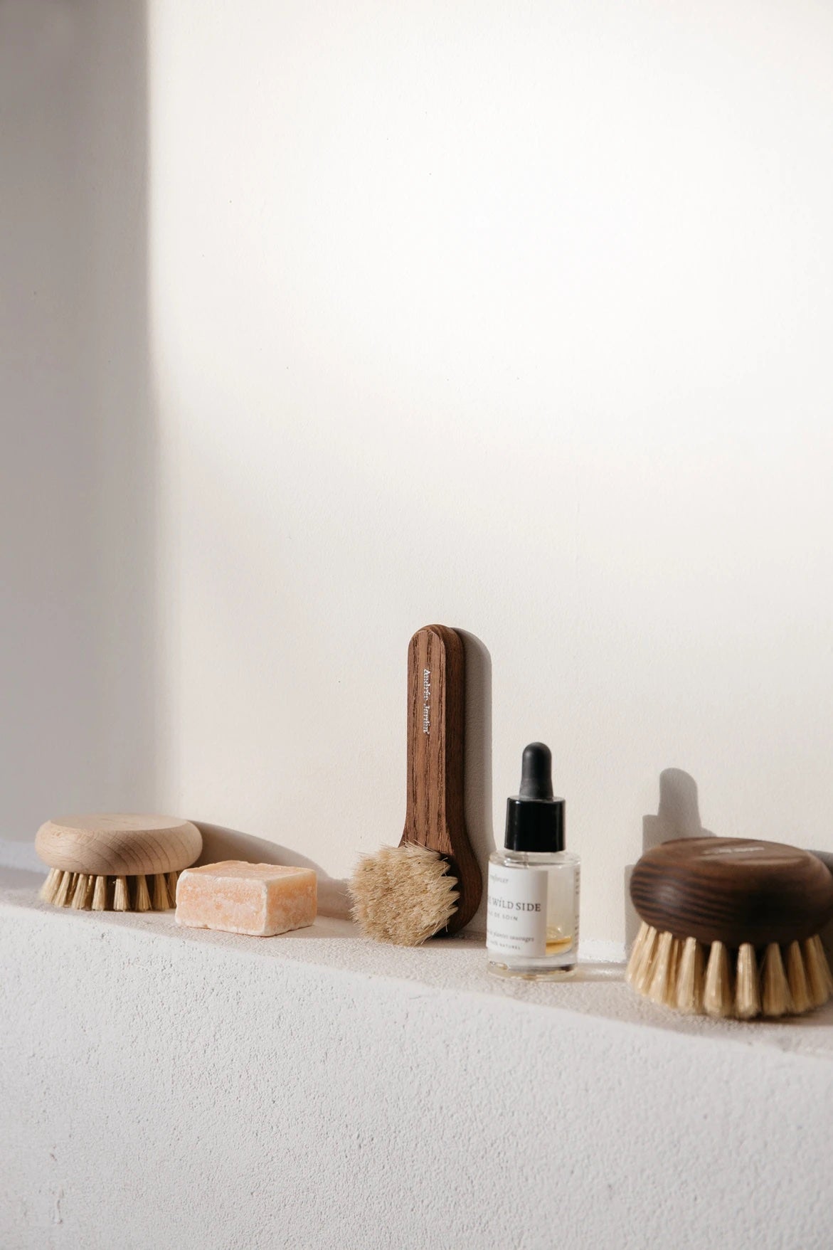 A collection of eco-friendly spa products, including Andrée Jardin Heritage Face Cleansing Brush Heat-Treated Ash Wood brushes and a bottle of oil, neatly arranged on a white ledge against a light gray wall.