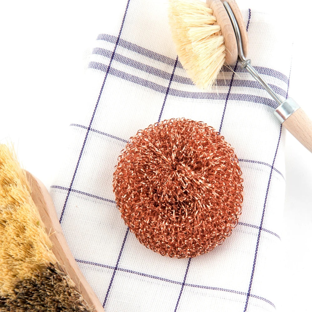 A Andrée Jardin Tradition Copper Scrubber rests on a blue and white checkered cloth, alongside a wooden brush with natural bristles, set against a white background.