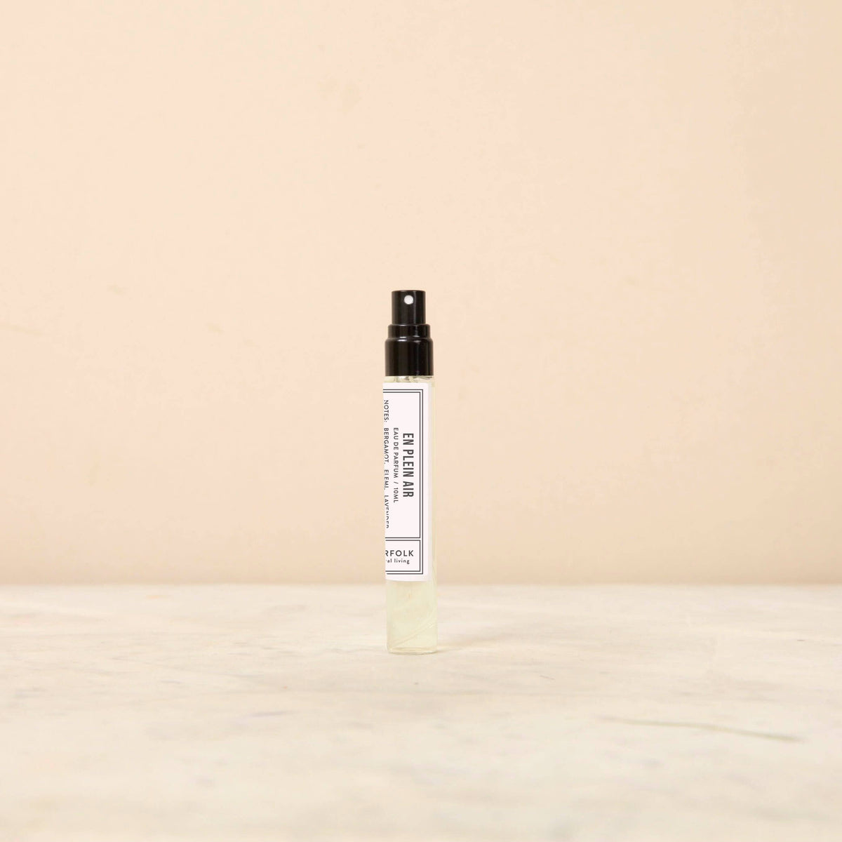 A small spray bottle with a black nozzle and cap stands on a light-colored surface against a beige background. The bottle has a white label with text and a minimalistic design, hinting at the clear liquid inside, which is infused with delicate floral essences. This product is the Norfolk Natural Living Parfum - En Plein Air 10ml by Norfolk Natural Living.