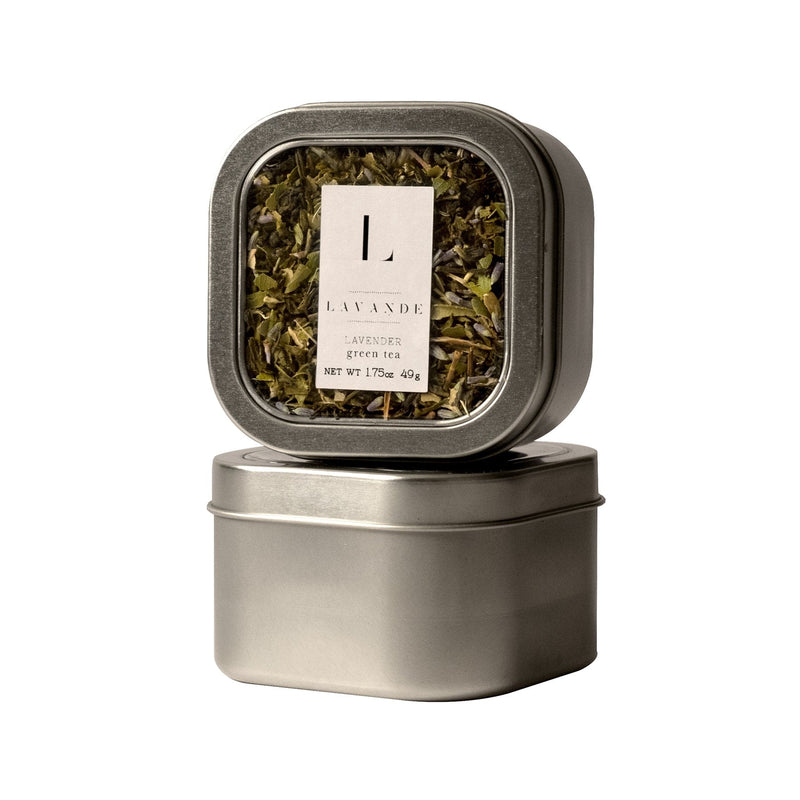 A metal tin with a transparent lid showcasing a new blend of Lavande - Lavender Lemon Ginseng Green Tea leaves, labeled "lavande lavender" with a minimalistic design. Another tin is stacked below it, facing forward.