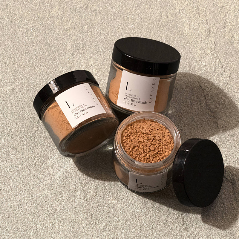 Three jars with labels, containing skincare products like Lavande - Lavender & Sandalwood Clay Face Mask and loose powder, positioned on a textured surface.