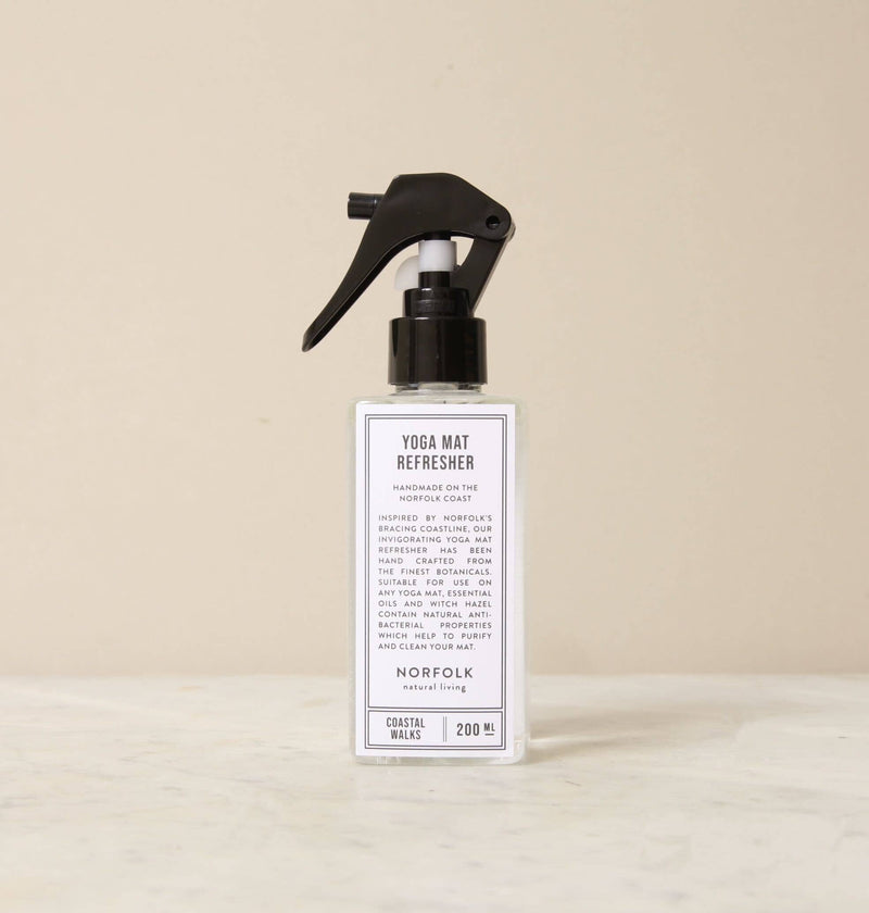 A spray bottle labeled "Norfolk Natural Living Coastal Yoga Mat Refresher" with anti-bacterial properties, standing on a neutral-colored countertop, with the focus on the clear label detailing the product's use and scent.