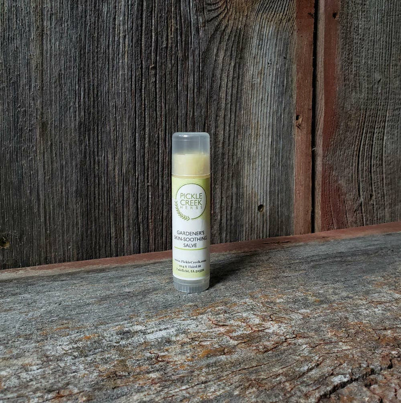 A tube of Pickle Creek Herbs Gardener's Skin-Soothing Salve stands upright on a rustic wooden surface, with weathered wood planks in the background providing a textured backdrop.