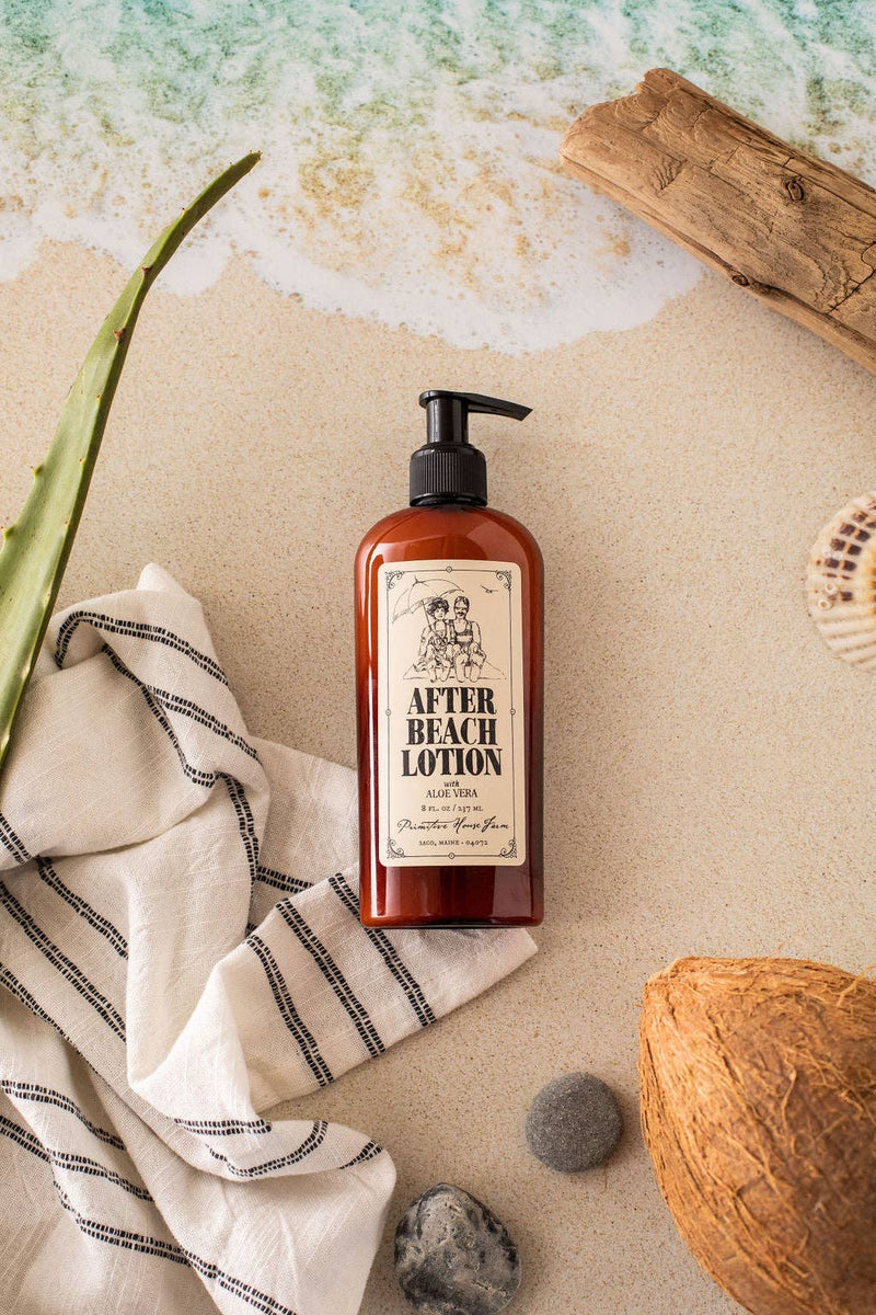A bottle of "Primitive House Farm - After Beach Lotion" is on a sandy surface with a towel, aloe vera, driftwood, and virgin coconut. The setting evokes a beach environment with an oceanic backdrop.