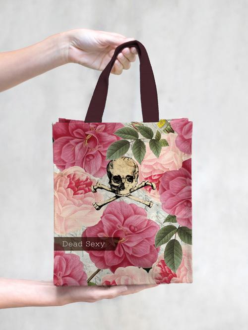 A hand holding a Margot Elena TokyoMilk Dead Sexy Small Tote adorned with a floral pattern, featuring pink blooms and leaves, and a distinctive skull and crossbones design with the phrase "dead sexy" across the center.