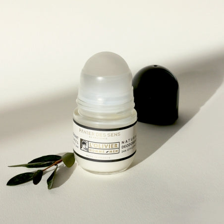A small, open jar of Panier des Sens L’Olivier Natural Deodorant beside its black cap, with a few green leaves on a sunlit white surface, utilizes.