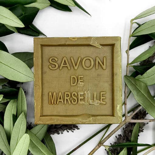 A bar of "Senteurs De France Marseille Raw Olive Oil Cube Soap" surrounded by fresh green leaves and small pine cones on a white background, highlighting the soap's natural ingredients.