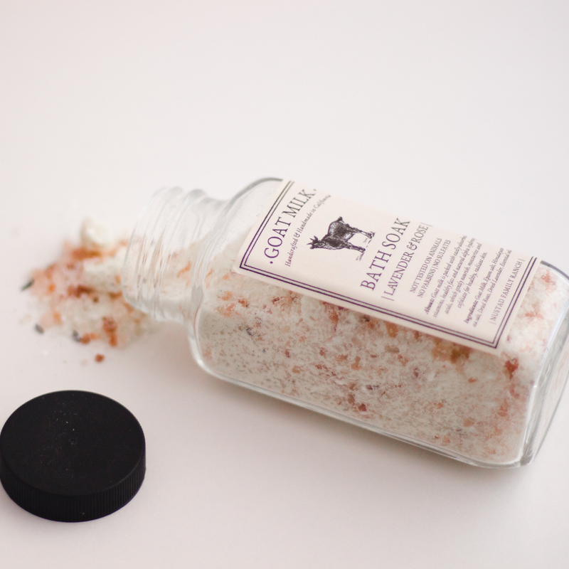 A clear glass bottle of Nustad Family Ranch Goat Milk Bath Soak tipped over with its black cap off and some salts scattered on a white surface.