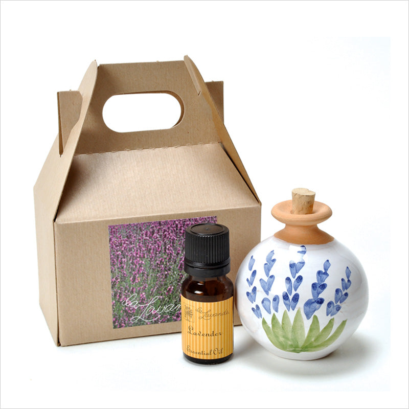 A brown cardboard carry box with a floral design next to a small brown glass bottle labeled lavender essential oil and a La Lavande Ceramic Lavender Oil Diffuser Gift Set hand-painted in Provence and a wooden