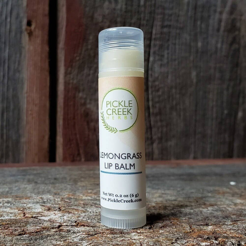 A tube of Pickle Creek Herbs Lemongrass Lip Balm stands on a wooden surface with a rustic wooden background. The label displays the product name and logo.