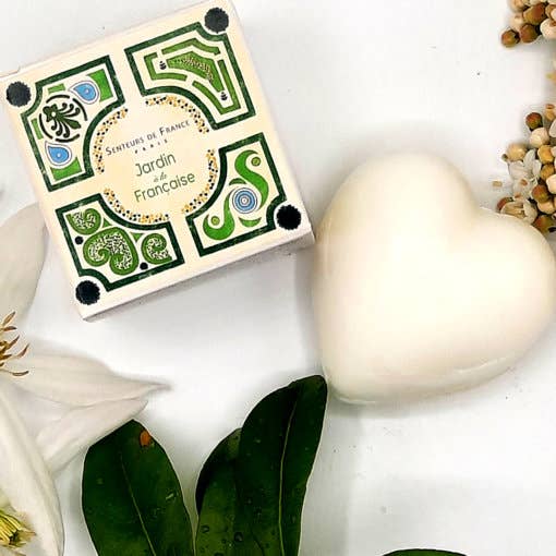 A Senteurs De France Lily of the Valley Heart Soap made with organic shea butter beside a square soap box with colorful, ornate patterns, surrounded by green leaves and small beads on a light background.