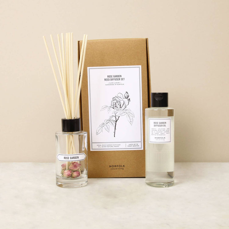 A Norfolk Natural Living Rose Garden Diffuser set displayed on a tabletop, including a glass bottle with reeds and pink petals, a large brown cardboard box with floral graphics, and a labeled clear bottle of English countryside aroma.
