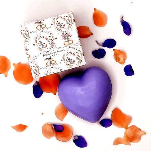 A purple, Senteurs De France Versailles lavender heart-shaped soap beside its decorative box, surrounded by scattered orange and blue flower petals on a white background.
