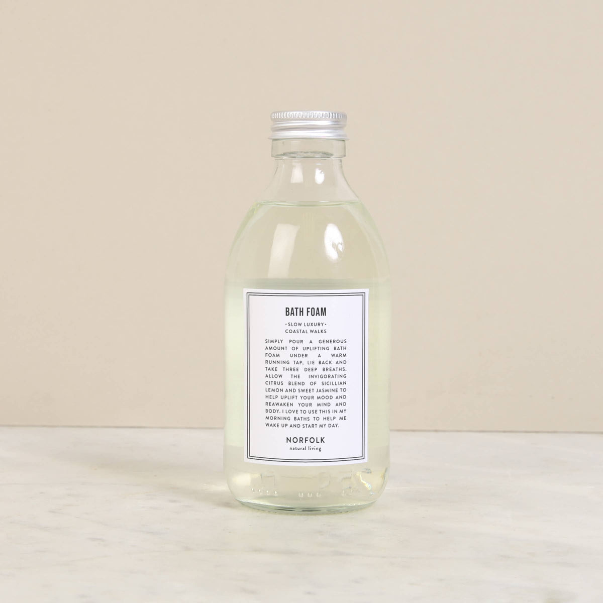 A clear glass bottle containing Norfolk Natural Living Coastal Bath Foam, 250ml infused with essential oils sits on a beige surface. It features a white label with black text that includes product details. The bottle has a silver screw cap.