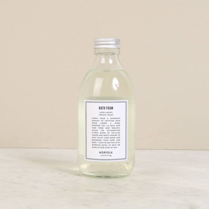 A clear glass bottle containing Norfolk Natural Living Coastal Bath Foam, 250ml infused with essential oils sits on a beige surface. It features a white label with black text that includes product details. The bottle has a silver screw cap.
