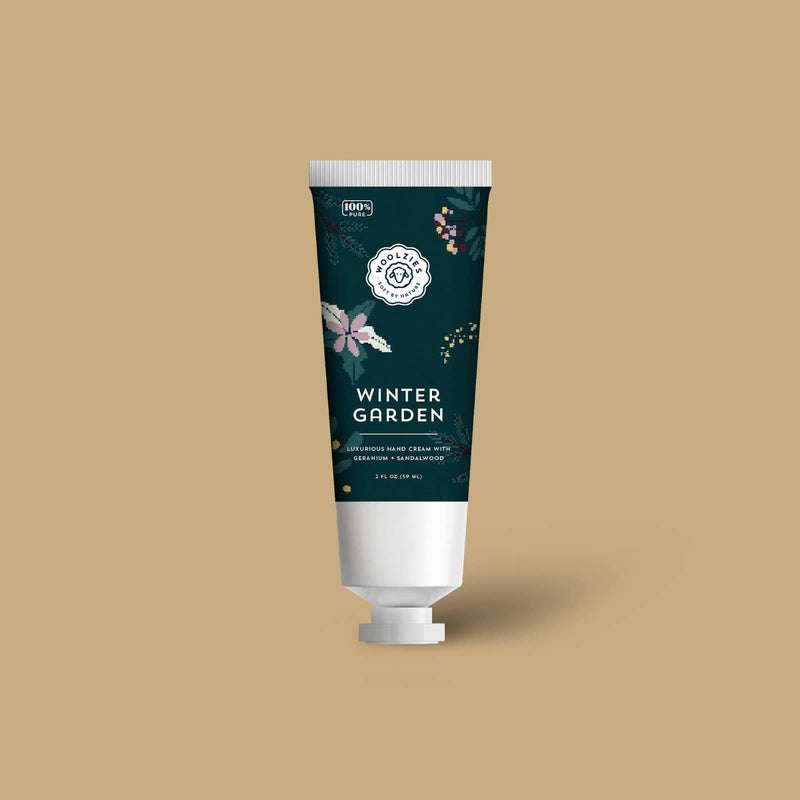 A tube of Woolzies Winter Garden Holiday Hand Cream 2oz enhanced with natural Shea butter on a plain beige background. The tube is decorated with floral and snowflake designs in dark tones with white and pink accents.