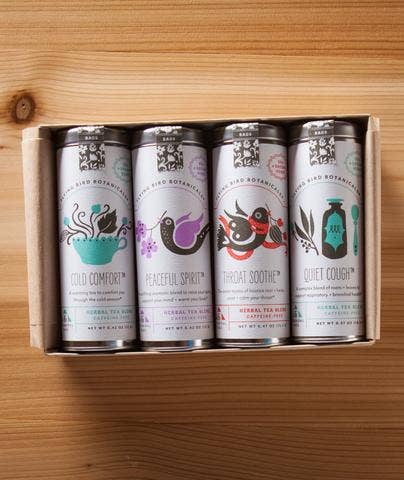 Four cans of Flying Bird Botanicals herbal tea in a biodegradable cardboard box, each labeled with different health benefits and adorned with colorful, whimsical artwork. The wood grain table provides a warm background.