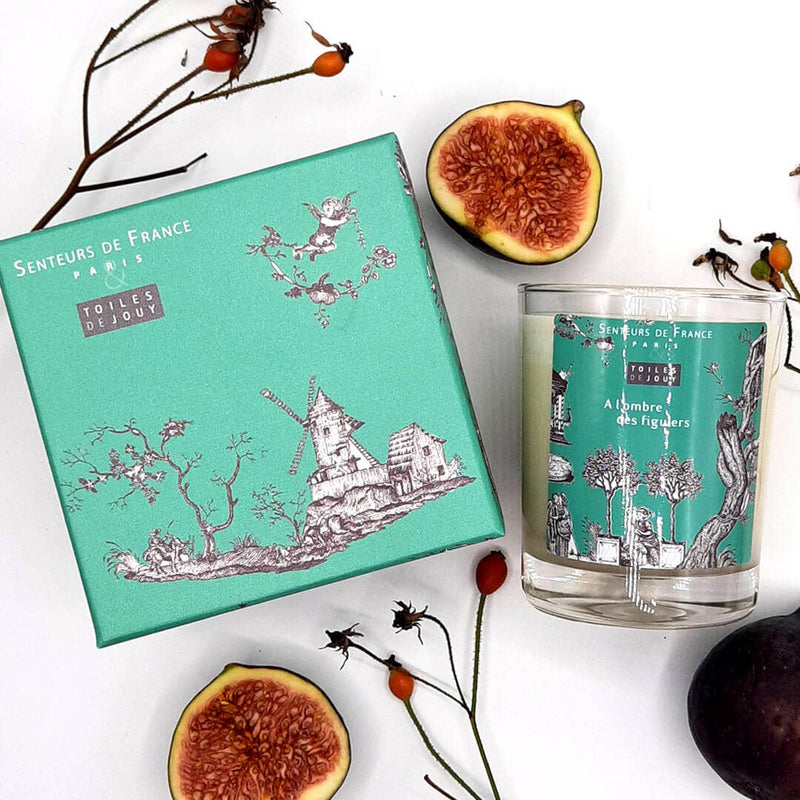 A flat lay image featuring a turquoise Senteurs De France Toiles de Jouy box labeled "senteurs de france" with intricate drawings, alongside a matching Senteurs De France scented candle and fresh figs on a white background.
