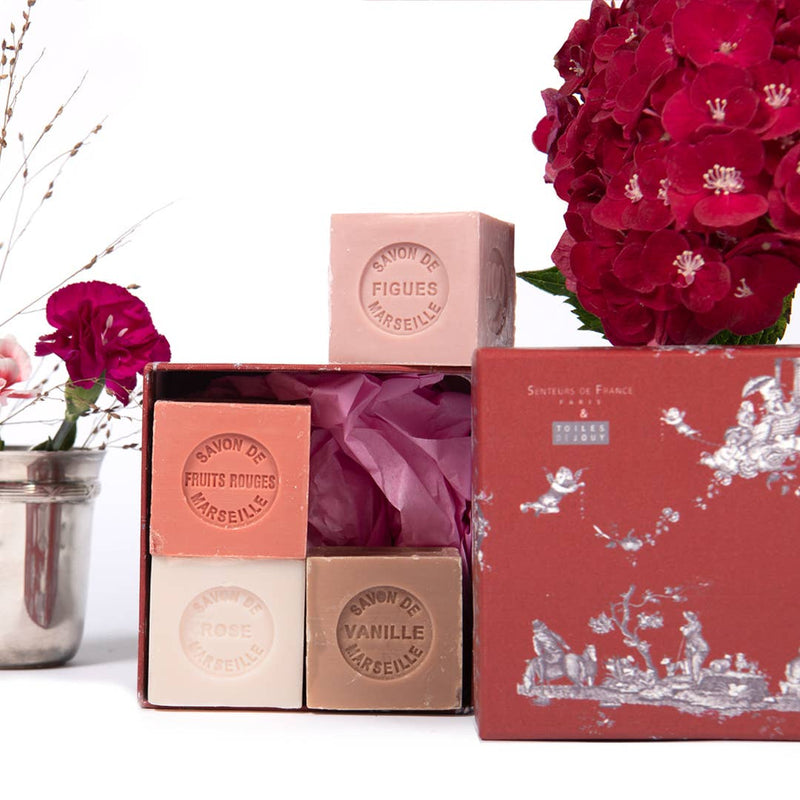 A display of Senteurs de France Coffret Vanilla, Red Fruits, Fig and Rose cube soaps next to a bouquet of red flowers. Four soaps labeled "fruits rouges," "rose," "vanille," and "figues de Marseille" are.