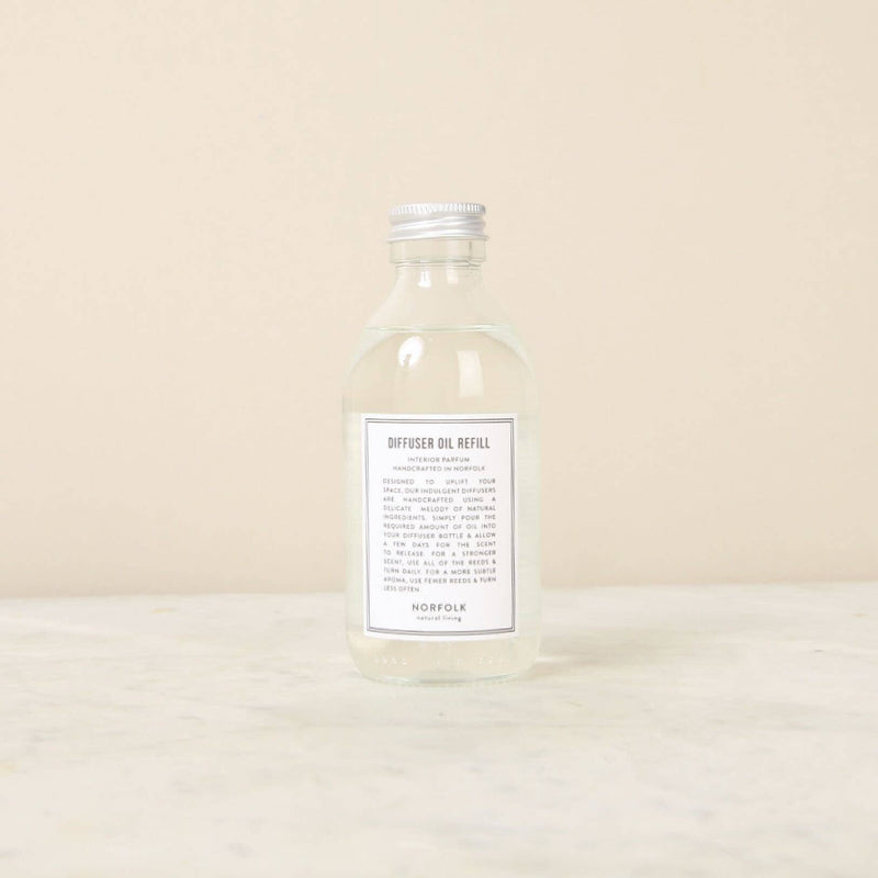 A clear glass bottle of Norfolk Natural Living Coastal Reed Diffuser Refill Oil with a white label, standing on a light beige surface against a matching background. The label reads "diffuser oil refill" along with other text details.