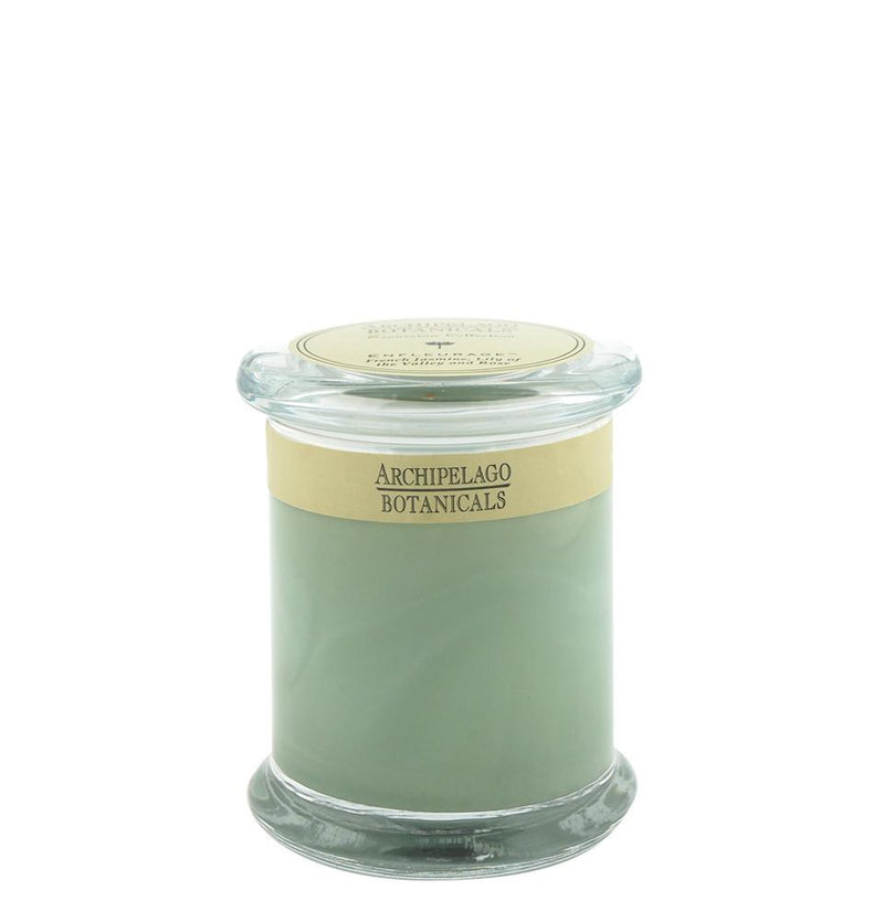 A glass jar with an Archipelago Excursion Enfleurage candle from Archipelago Botanicals, featuring a golden label around the middle, set against a white background.