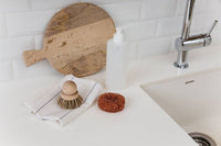 A kitchen countertop with a wooden cutting board, a white soap dispenser, a scrub brush on a cloth, and an Andrée Jardin Tradition Copper Scrubber near a sink and faucet.
