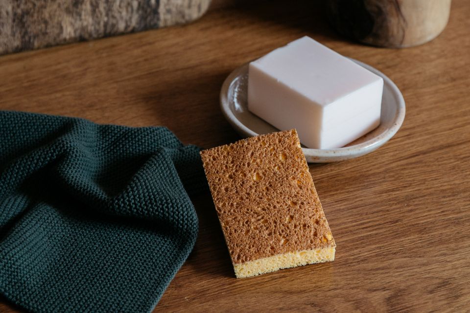 A biodegradable Andrée Jardin Natural Dish Sponge (set of 2) and a white bar of soap on a small ceramic dish, placed next to a dark green cloth on a wooden surface.