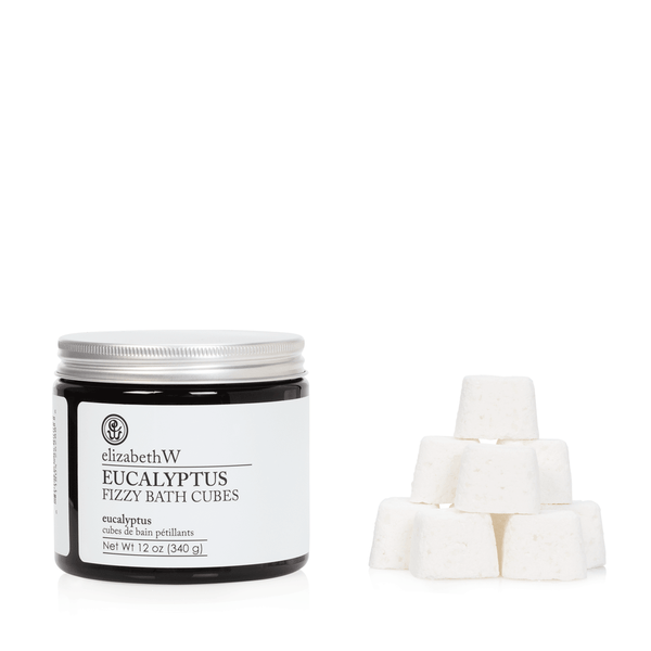 A jar labeled "elizabeth W Purely Essential Eucalyptus Fizzy Bath Cubes" next to a stack of six white, cube-shaped bath fizzies, isolated on a white background.