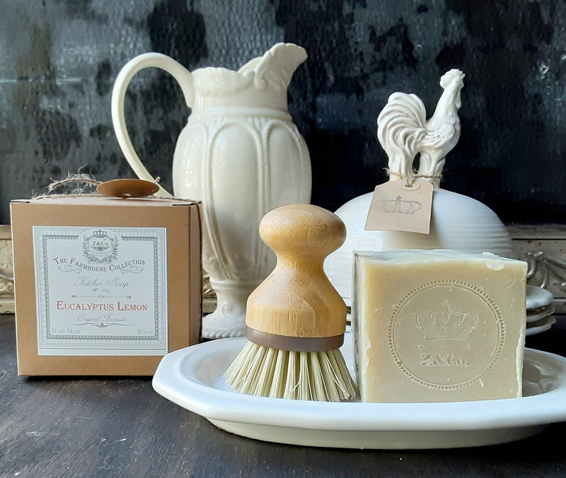 A rustic display of bath products including Z&Co. Farmhouse Solid-Block Eucalyptus Lemon Dish Soap, a wooden scrubbing brush, and a ceramic rooster-shaped jug, set against a dark background.