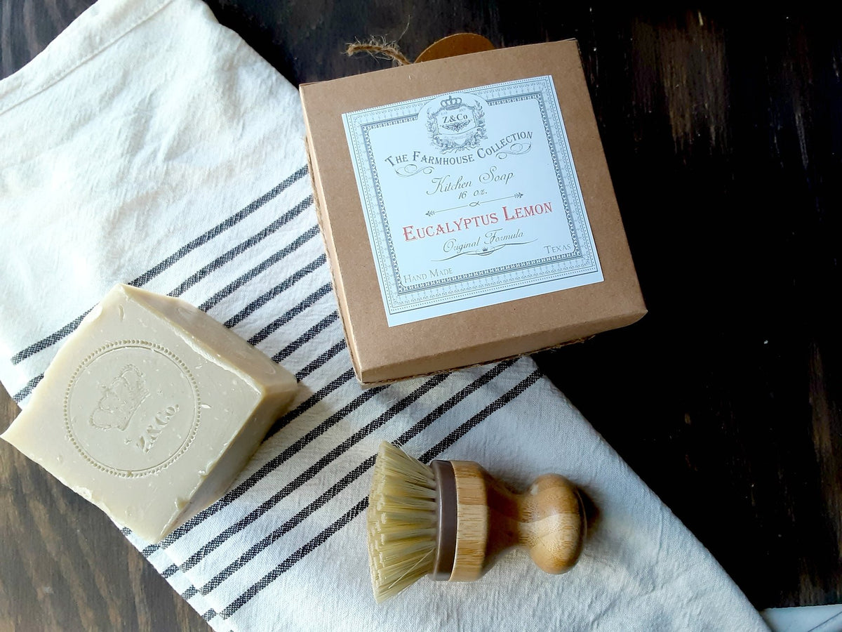 Z&Co. Farmhouse Solid-Block Eucalyptus Lemon Dish Soap bar next to its packaging and a wooden saucepan brush on a striped towel and dark wood surface.