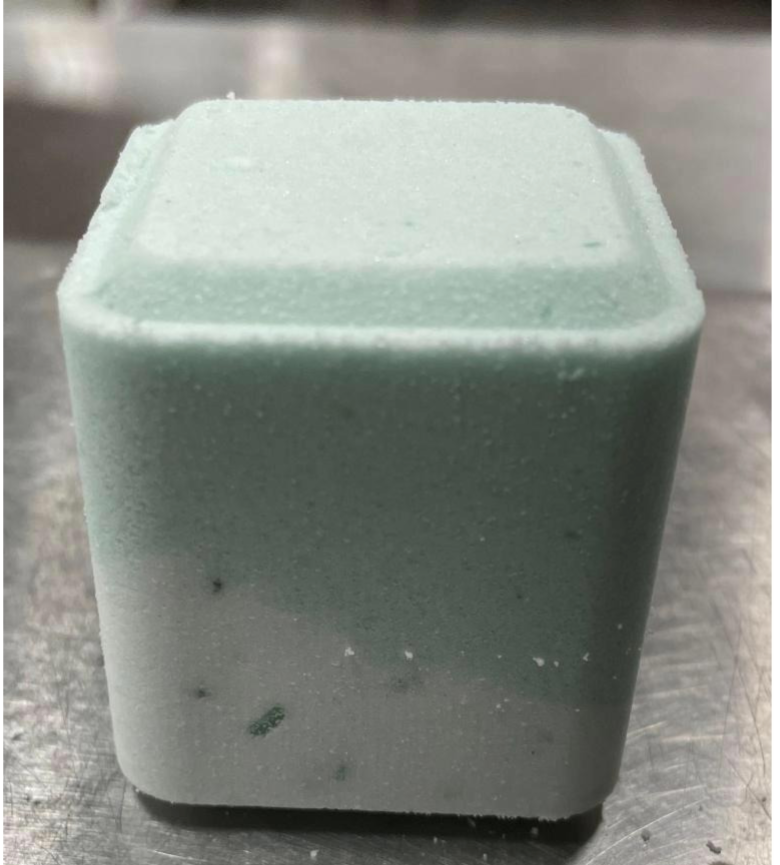 A close-up image of a Lizush Mint Shower Steamer, a pale blue, square-shaped block of shower steamer on a metal surface, showing minor visible particles and slight imperfections on its surface.