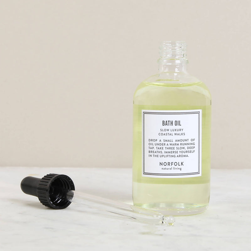 A clear bottle of light yellow Norfolk Natural Living Coastal Bath & Massage Oil, 100ml with a black cap beside it, labelled "slow luxury bath oil," placed on a marble surface. A glass dropper with some essential oils is lying in front.