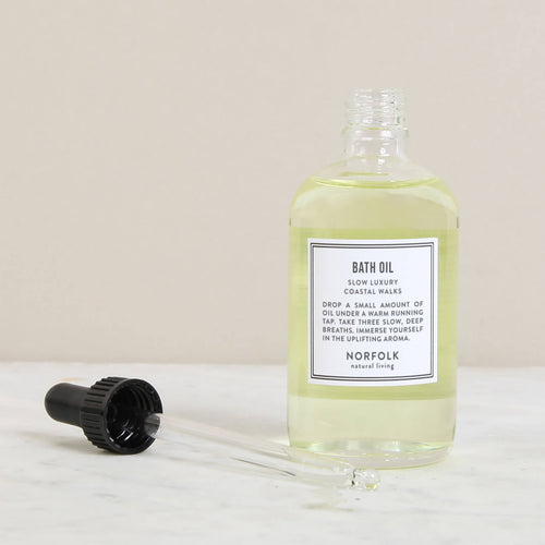 A clear bottle of light yellow Norfolk Natural Living Coastal Bath & Massage Oil, 100ml with a black cap beside it, labelled "slow luxury bath oil," placed on a marble surface. A glass dropper with some essential oils is lying in front.