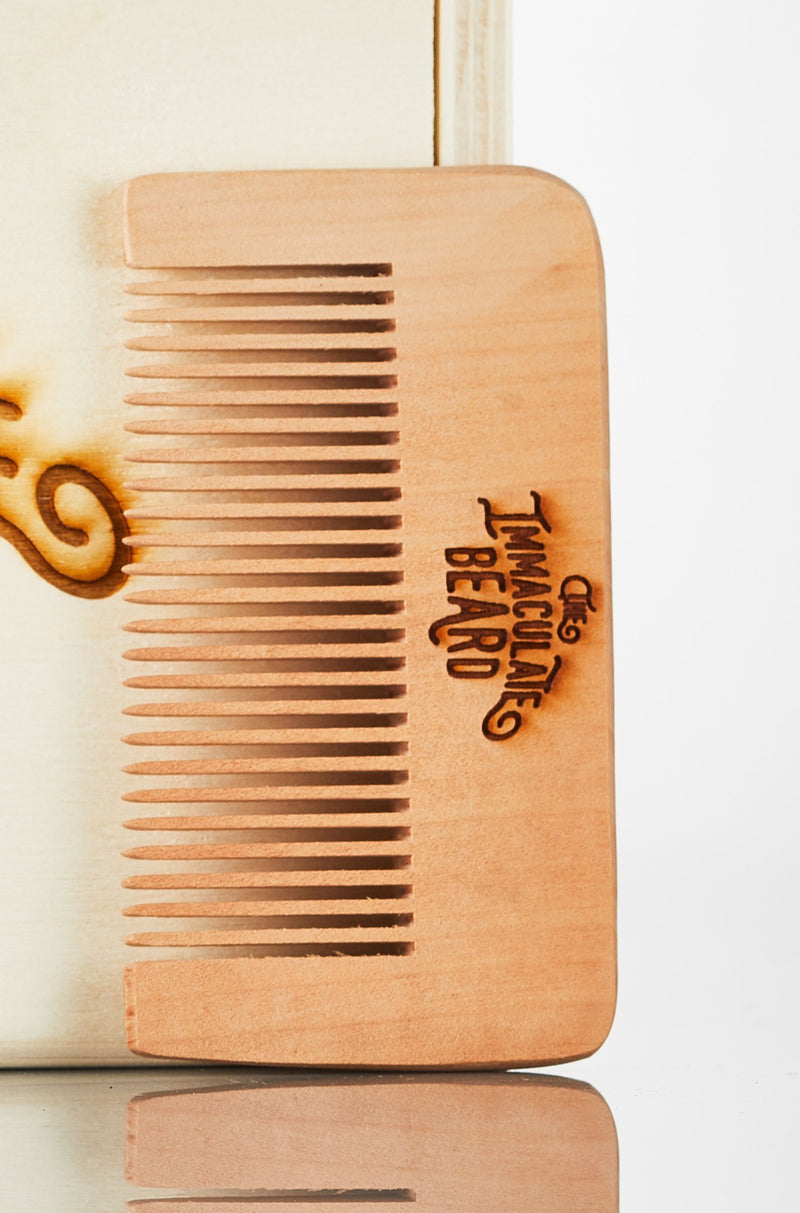 Close-up of The Immaculate Beard - Wooden Beard Comb with fine and wide teeth, featuring the engraved text "taming the beard" on its body, placed against a light background.