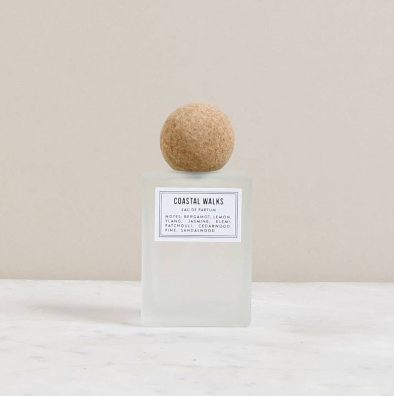 A minimalist unisex Norfolk Natural Living parfum bottle with a square base and a spherical cork stopper. The label reads "coastal walks" with scent notes of bergamot, lemon, and sand.